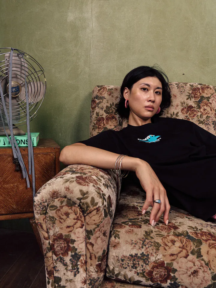 Woman in a black Coddiwomple Studio t-shirt leaning on the arm of a floral sofa next to a vintage fan