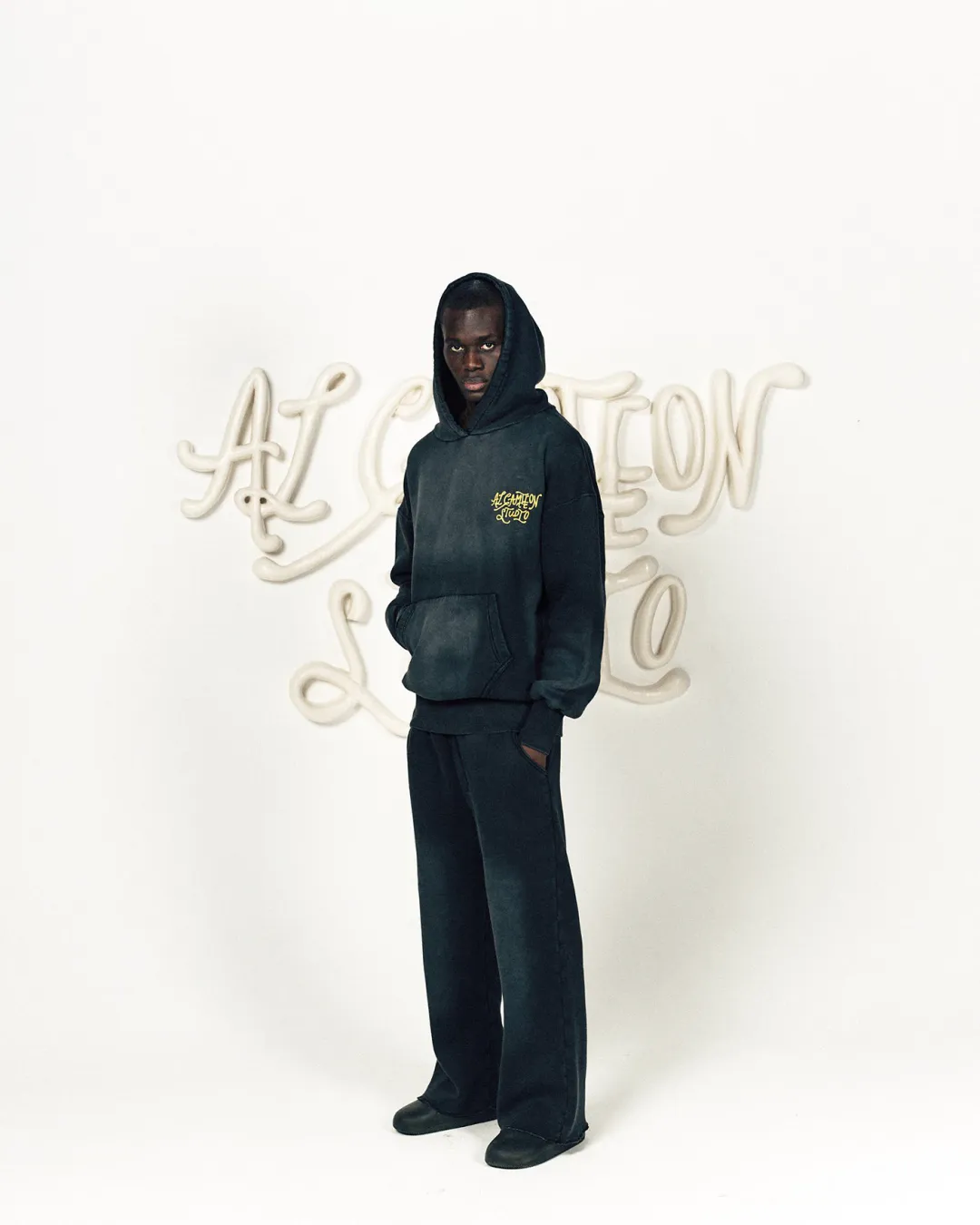 Man in a dark washed hoodie standing in front of an Al Camileon Studio backdrop