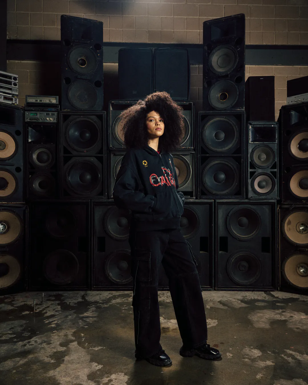 Woman with voluminous curly hair in a dark hoodie in front of a wall of speakers