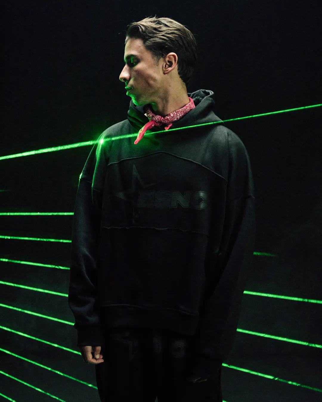 Man in a black hoodie and red bandana in profile, lit by green laser beams