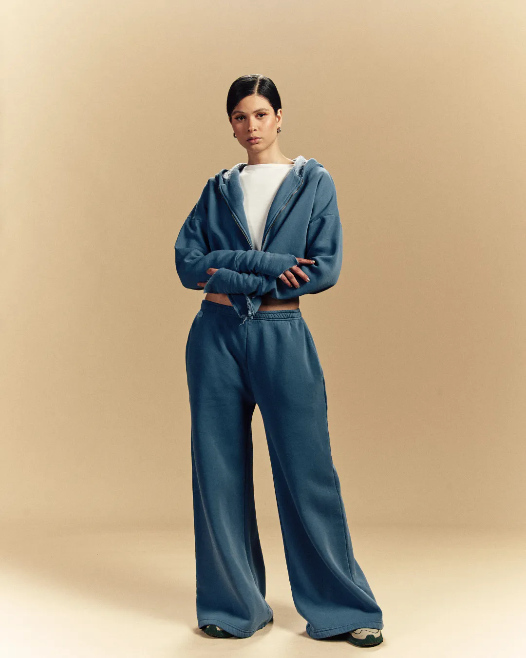 Model in a blue cropped hoodie and wide-leg sweatpants against a warm beige studio backdrop