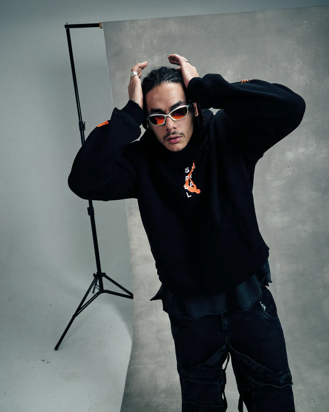 Man in sunglasses and a black hoodie with both hands on his head against a grey studio backdrop