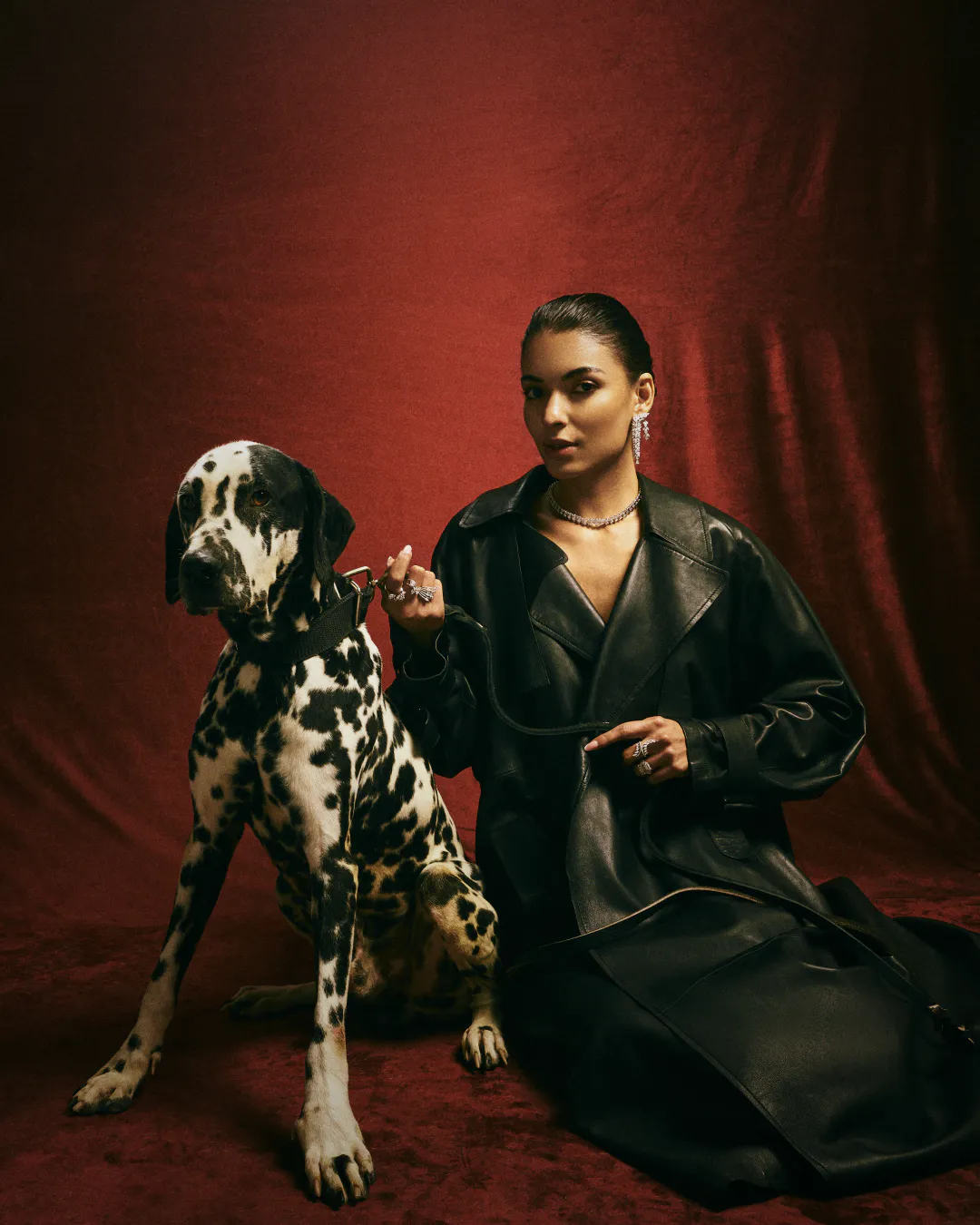 Woman in a black leather coat seated beside a Dalmatian against a deep red backdrop
